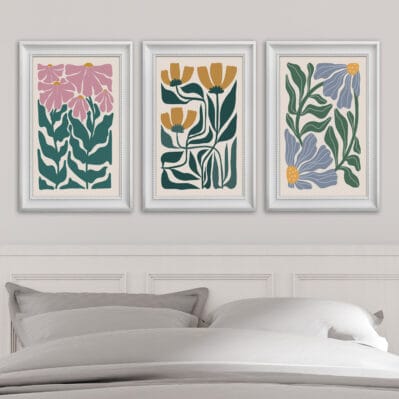 Set di tre quadri decorativi per testata letto con illustrazioni floreali e cornici stampate, appesi sopra un letto matrimoniale.